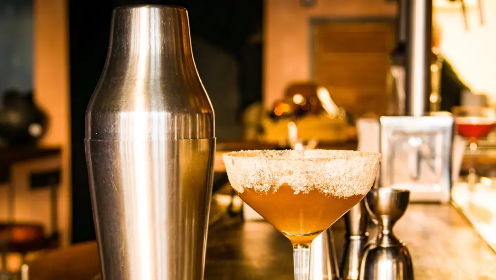 Close up image of a French cocktail shaker sat next to a cocktail in a bar room setting.