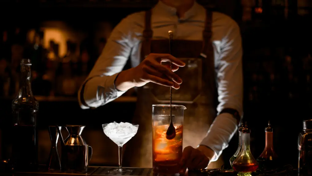 image of darkly lit bar with bartender using a bar spoon while mixing a drink