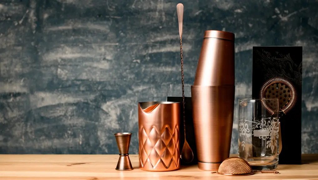 Close up image of bar tools a jigger, a shaker and a strainer in a bronze metalic colour.