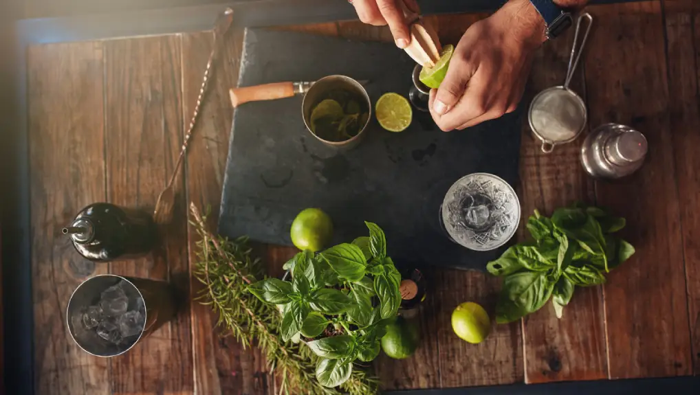 overhead image of a bartender preparing a cocktail on a bar top with a selection of  limes, mint and bar tools