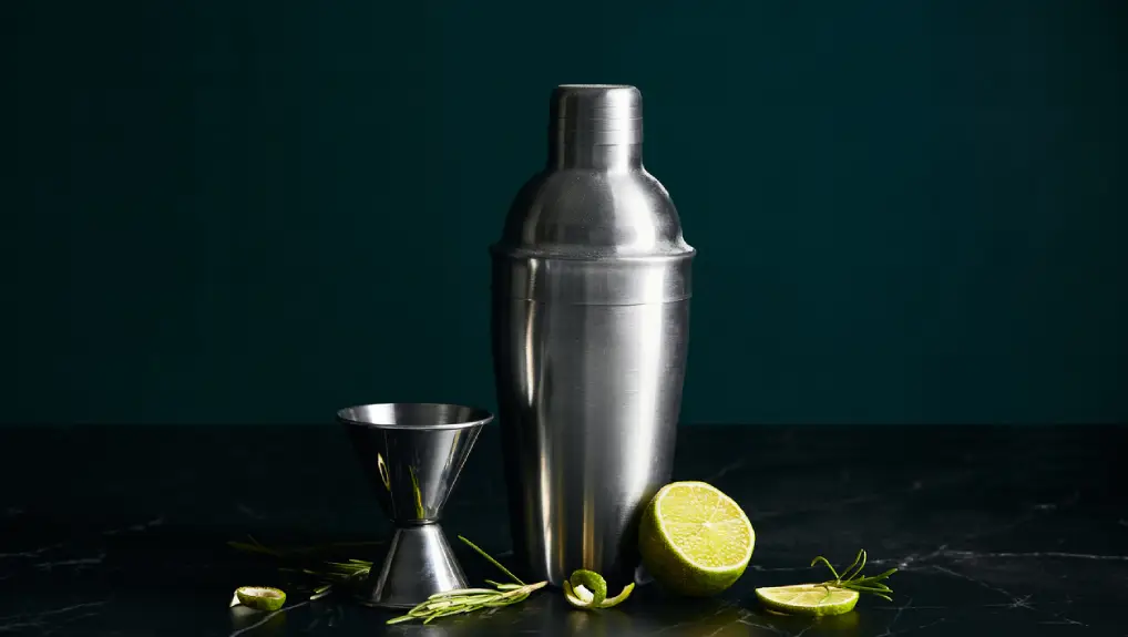 close up imager of a silver cocktail shaker, a jigger and a lime. the image has a dark green background