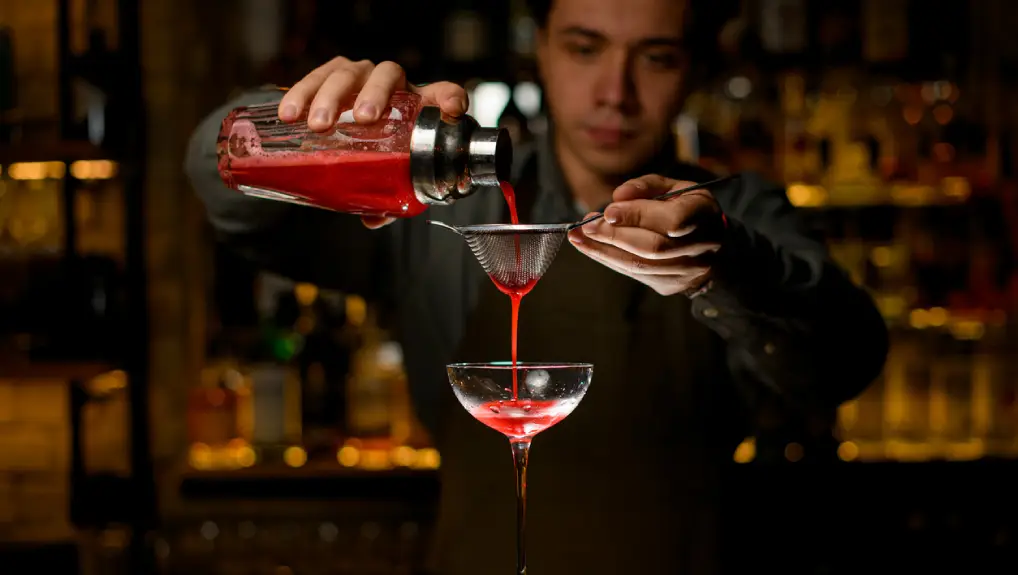 Close up image of a bartender pouring from a vintage Boston cocktail shaker in a trendy bar the lighting is dark and casting a deep red colour